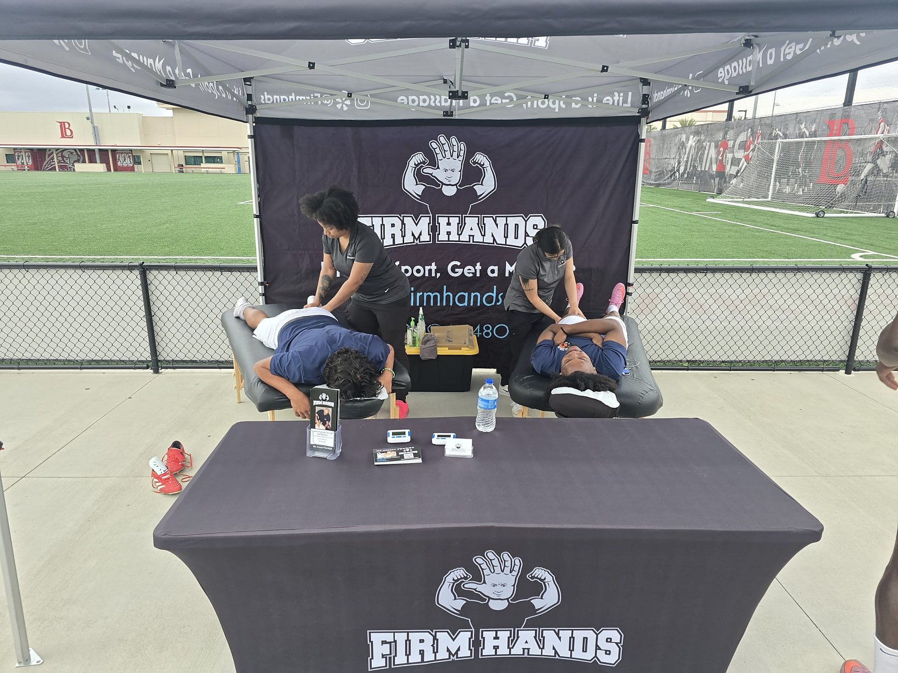 Two athletes on adjacent tables under the Firm Hands banner at a school field